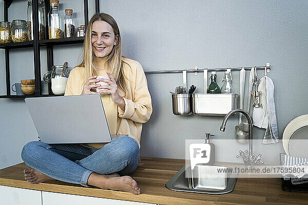 Junge Frau mit Kaffeetasse und Laptop sitzt auf der Küchentheke