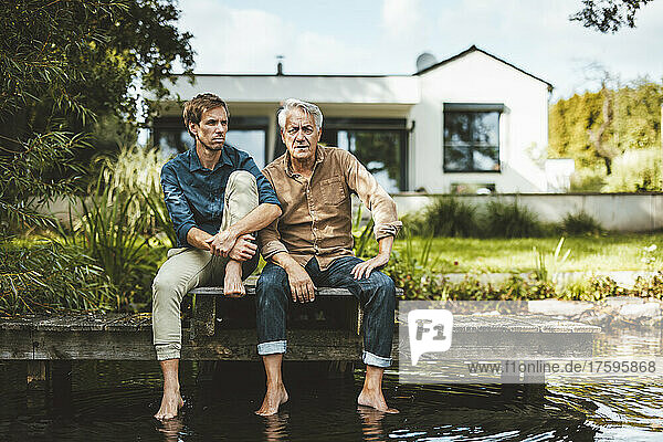 Senior man with son sitting on jetty by lake at backyard