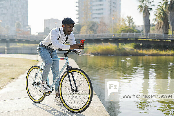 Young man using smart phone on bicycle by lake in city