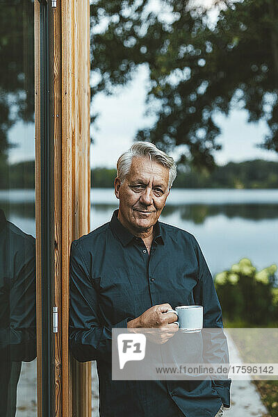Senior man with coffee cup standing by glass wall at backyard