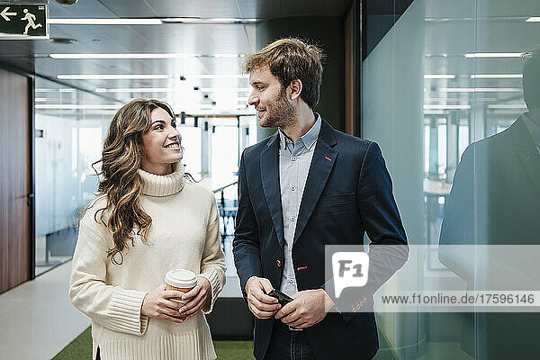 Smiling businessman and businesswoman looking at each other by glass wall in office