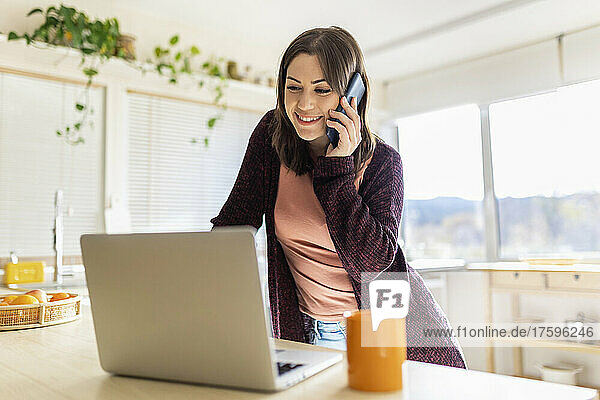 Young businesswoman talking on smart phone by laptop in kitchen at home