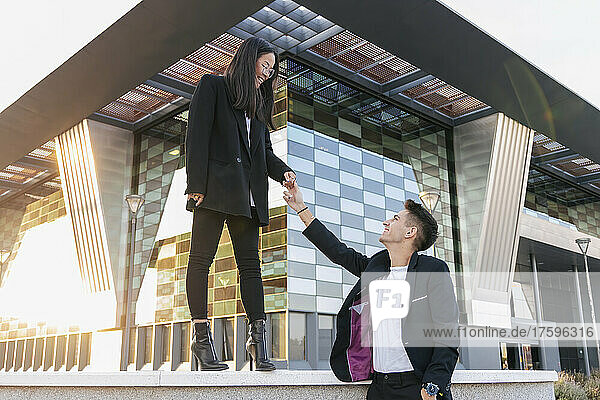 Smiling businessman holding hand of colleague standing on wall in front of office building