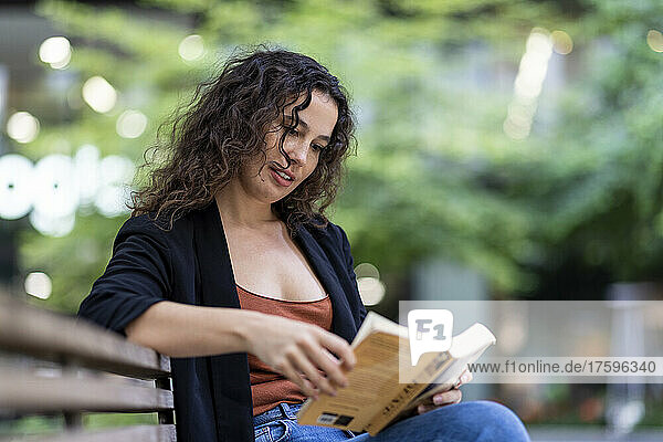 Young woman reading book on bench in park