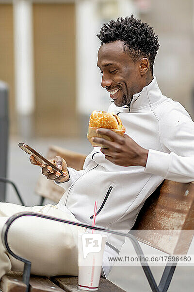 Smiling man holding burger using mobile phone sitting on bench