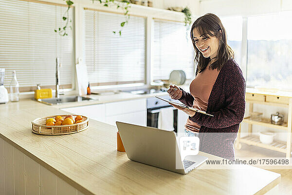 Smiling businesswoman with clipboard looking at laptop on table at home