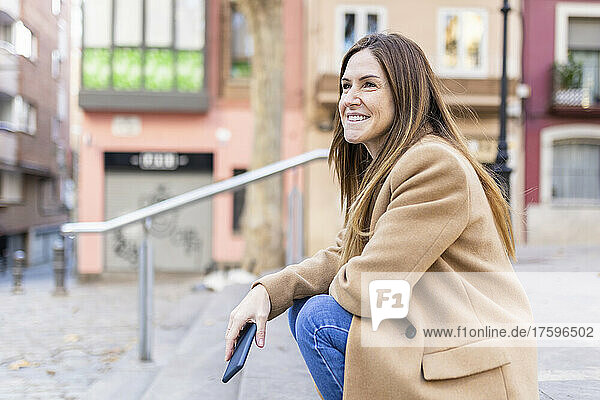 Smiling woman with smart phone sitting on footpath in city