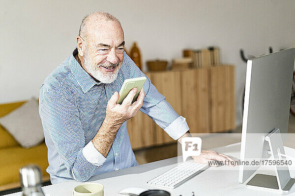 Smiling senior businessman sending voicemail through smart phone at desk