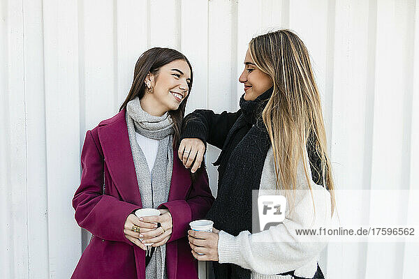 Young friends holding disposable cups smiling in front of white corrugated wall