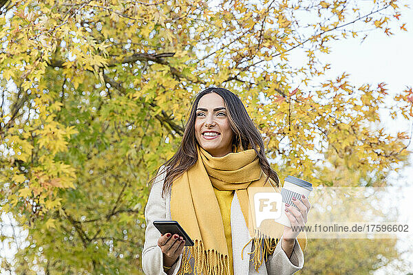 Smiling young woman with smart phone and reusable coffee cup in autumn park