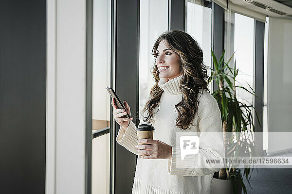 Lächelnde Geschäftsfrau mit Smartphone hält Einwegbecher am Fenster im Büro