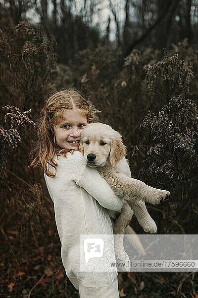 Smiling girl carrying golden retriever puppy standing in front of tree