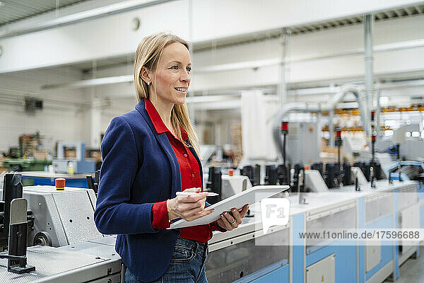Smiling blond businesswoman with tablet PC standing by machinery in factory