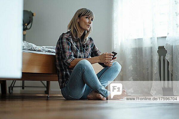 Contemplative woman with mobile phone sitting in bedroom at home