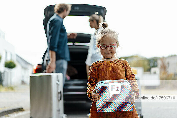 Girl holding suitcase with parents loading luggage in car
