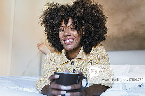 Smiling woman with coffee cup lying on bed in bedroom