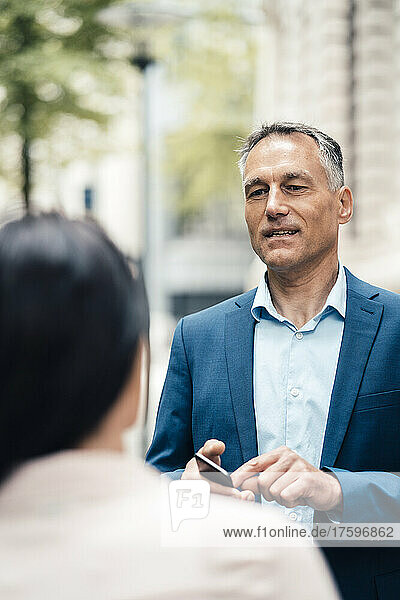 Mature businessman holding smart phone talking with businesswoman