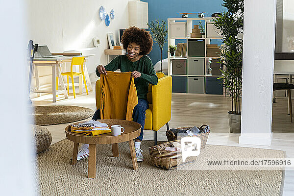 Smiling young Afro woman folding t-shirt sitting on chair at home