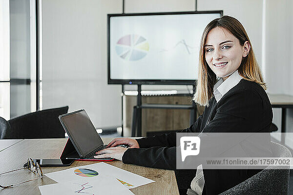 Smiling young businesswoman with tablet PC sitting at desk in office