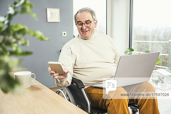 Smiling senior disabled man checking mobile phone sitting on wheelchair at home