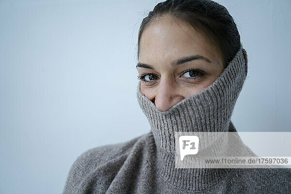 Playful woman hiding face with gray sweater in front of wall