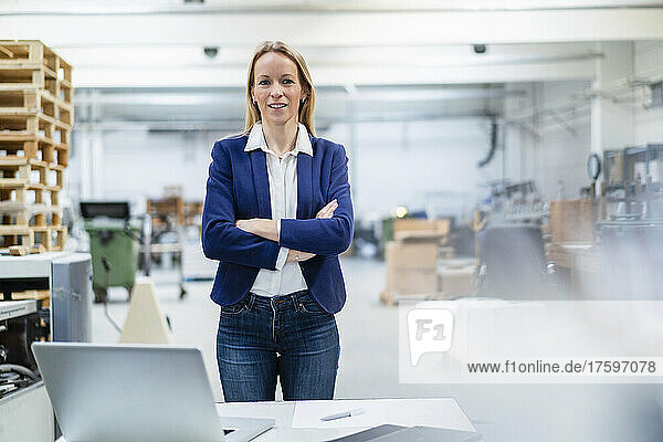 Blond businesswoman standing with arms crossed at desk in factory