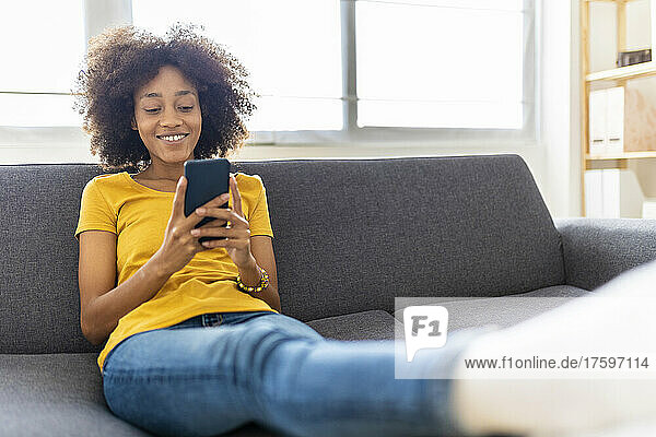 Smiling young woman surfing net through smart phone on sofa at home