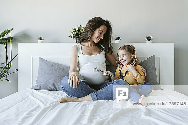 Smiling pregnant mother looking at daughter using stethoscope on bed