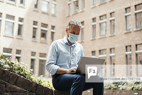 Businessman in protective face mask using laptop at office park
