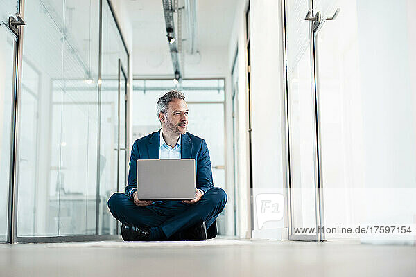 Thoughtful businessman sitting cross-legged with laptop in office corridor