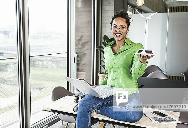 Smiling businesswoman with wind turbine model and laptop in office
