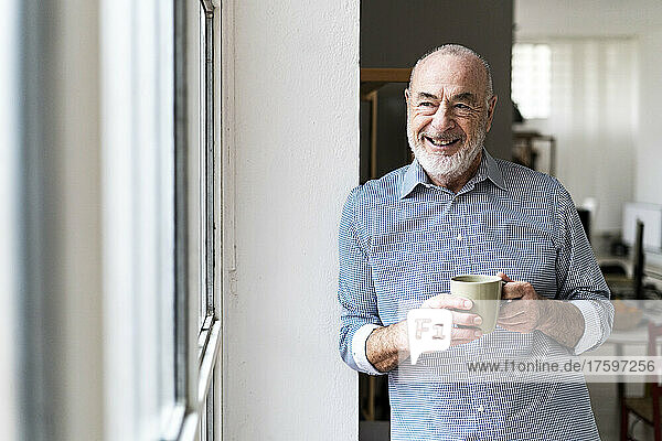 Happy senior freelancer holding coffee cup looking through window