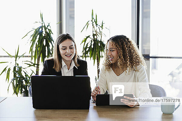 Young businesswoman with tablet PC looking at colleague using laptop at desk in coworking office