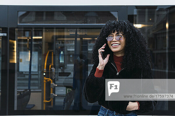 Happy young woman talking on smart phone in front of tram