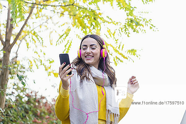 Smiling young woman enjoying music through headphones in autumn park