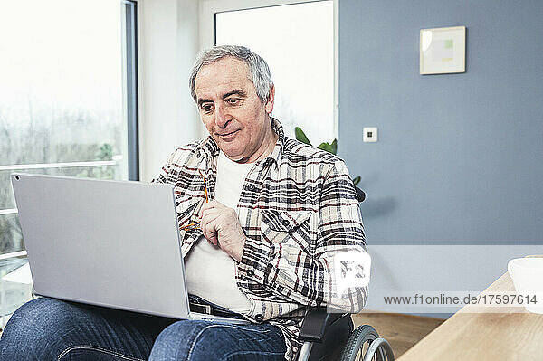 Disabled senior man with laptop sitting on wheelchair at home