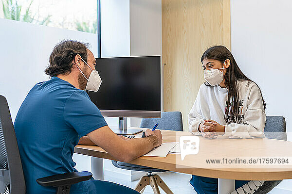 Doctor discussing with patient sitting at desk in hospital during pandemic