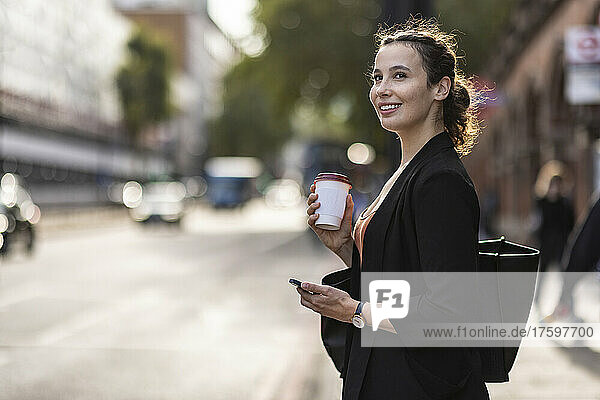 Thoughtful businesswoman holding smart phone and disposable coffee cup on street