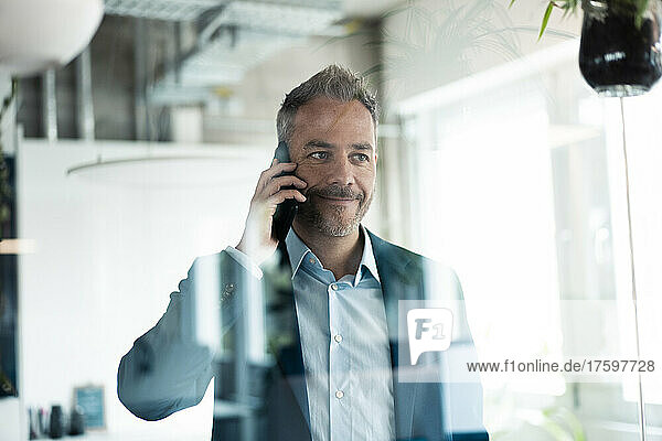 Smiling businessman looking away talking on mobile phone seen through glass wall