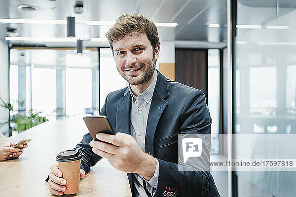 Lächelnder Geschäftsmann mit Einwegkaffeetasse und Smartphone am Schreibtisch