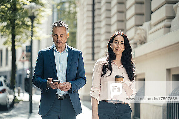 Businesswoman with disposable coffee cup walking by businessman using smart phone on footpath