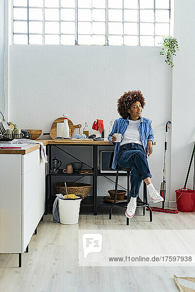Nachdenkliche junge Afro-Frau sitzt auf einem Hocker und lehnt an der Küchentheke und entspannt sich zu Hause