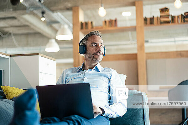 Businessman listening music through wireless headphones sitting with laptop on sofa at office