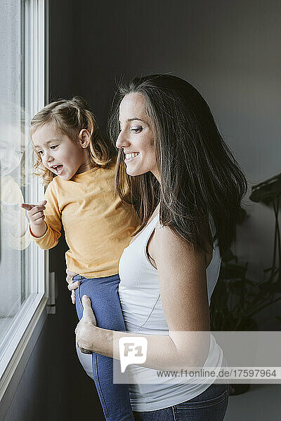 Happy pregnant woman with daughter looking through window at home