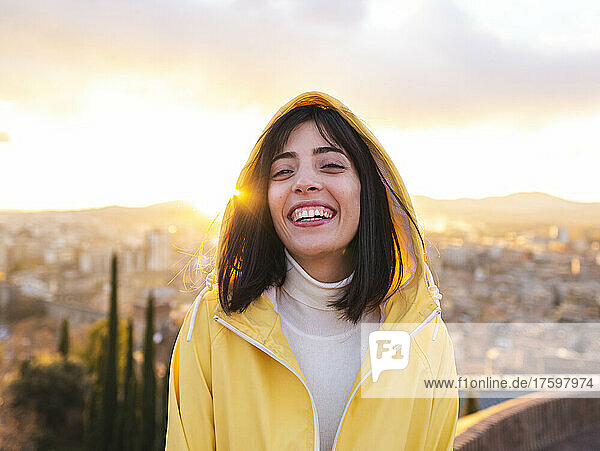 Lächelnde junge Frau im gelben Regenmantel mit Stadt im Hintergrund bei Sonnenuntergang