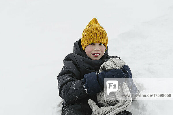 Boy wearing yellow knit hat holding gray scarf sitting on snow