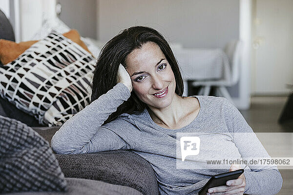 Lächelnde Frau mit Smartphone lehnt zu Hause auf dem Sofa