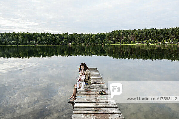 Mother spending leisure time with baby girl by lake on jetty