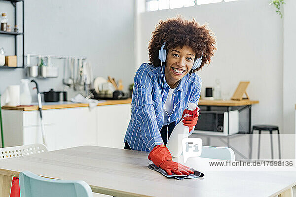 Smiling Afro woman listening music cleaning dining table in kitchen