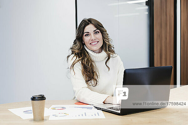 Lächelnde Geschäftsfrau sitzt am Schreibtisch mit Laptop im Büro